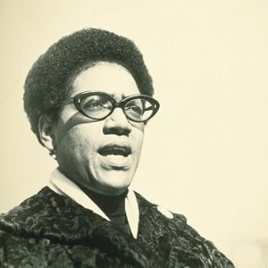 A black-and-white image of Audre Lorde speaking, wearing her signature glasses.