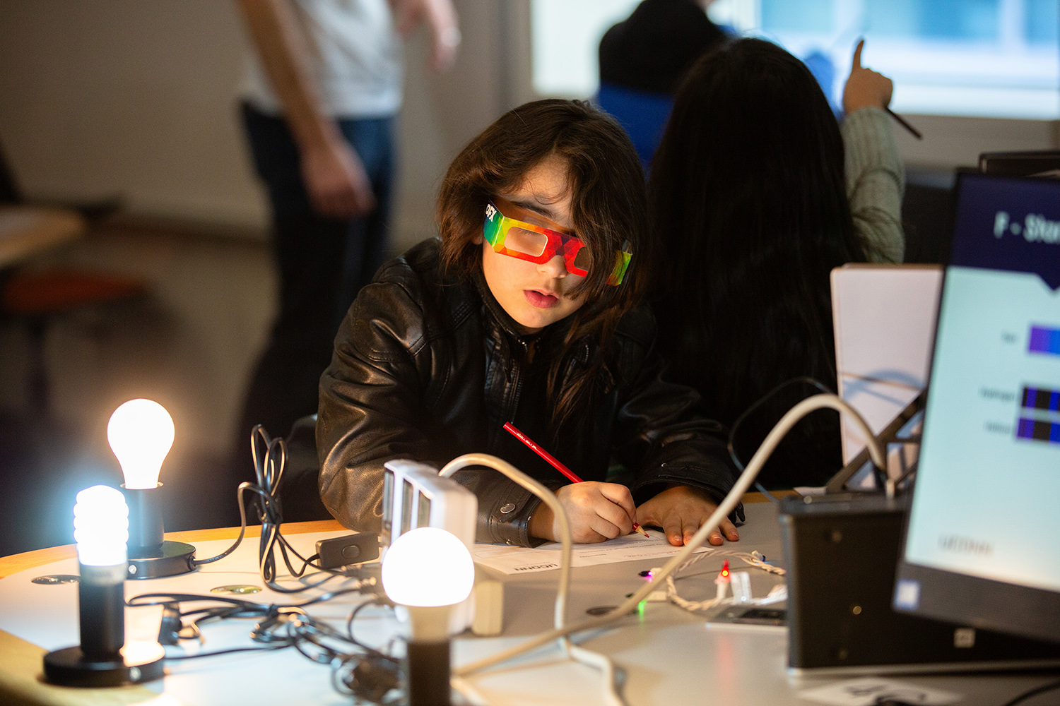 A student wearing glasses writes on a piece of paper during a physics outreach event.
