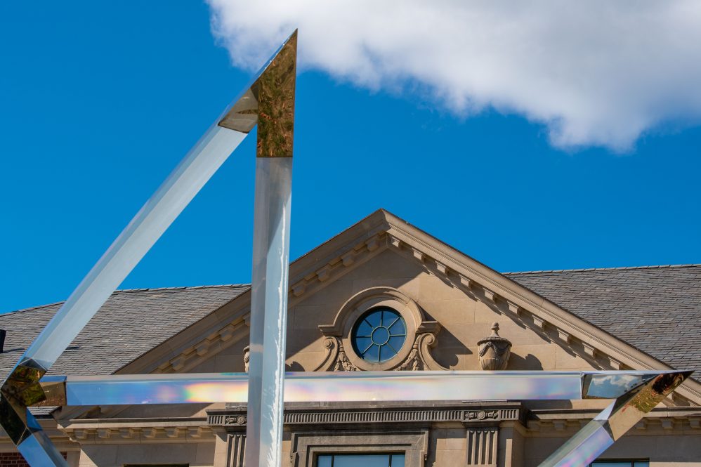 An angular prism structure reflects rainbows in front of an academic building and a blue sky.