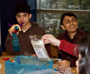 Two male college students sit at a table. One looks at the camera while holding a sock puppet.