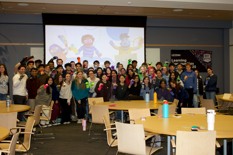 A group of college students poses for a photo in a classroom while wearing sock puppets on their hands.