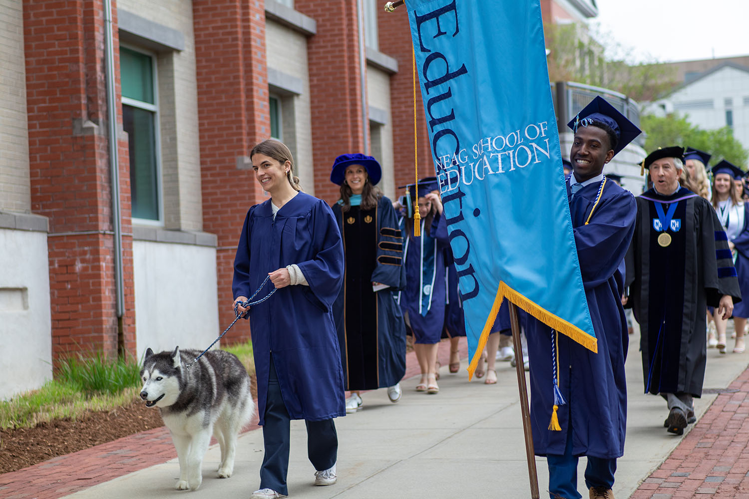 Neag School Class of 2024 graduates process alongside the Gentry Building led by a Black male student carrying the School's light blue banner.