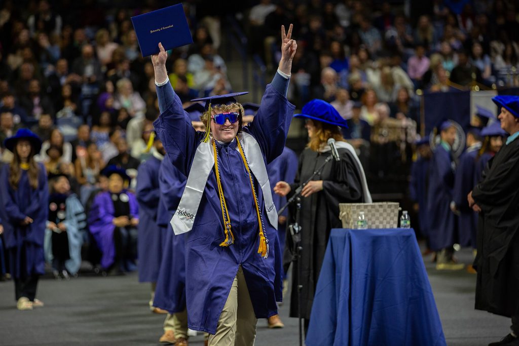 Student throws peace sign in air while walking across floor during commencement ceremony.