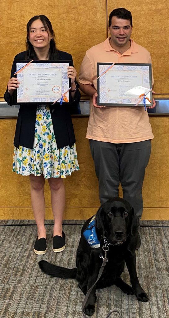 two graduates holding certificates and a black lab