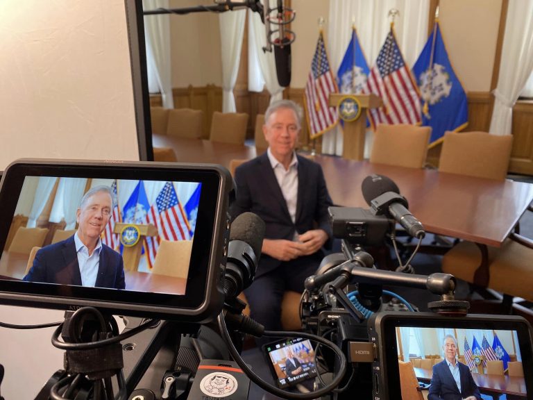 CT Governor Ned Lamont sits in front of US and Connecticut flags, being filmed for the documentary.