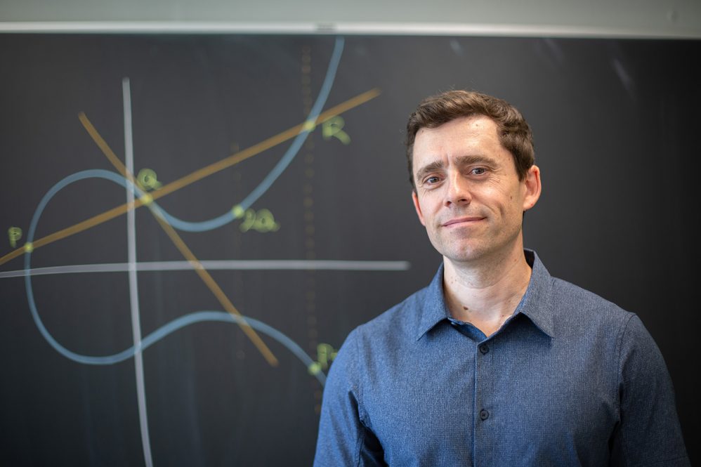 Alvaro Lozano-Robledo stands in front of a blackboard that has an equation written on it.