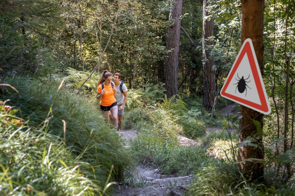 A woman and man hike through woods, with a warning sign about ticks on the trail ahead of them.