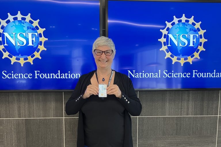 Marie Coppola smiling, holding up her badge in front of NSF logo.