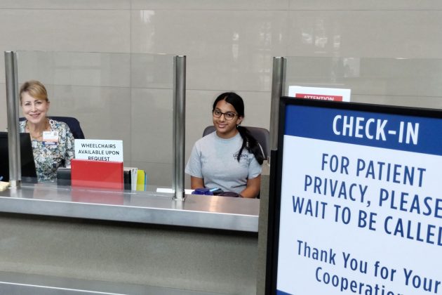 Portrait of two women at hospital information desk with check-in sign in foreground