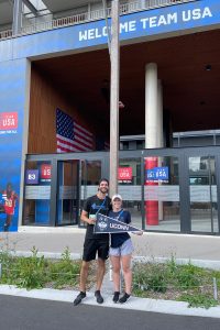 Two individuals, one male and one female, hold a UConn pennant while standing in front of the Team USA headquarters at the Paris Olympics.