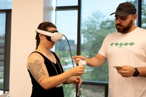 An adult man guides an adult woman who is wearing a VR headset.