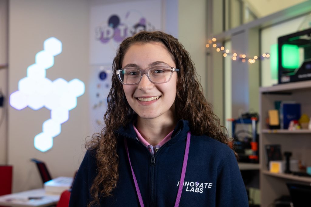 Sophia Hatzis, wearing glasses and a purple lanyard, poses for a photo in front of a background with colored lights.