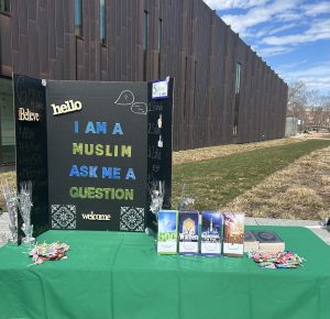 A table covered with a green tablecloth. It holds informative brochures and a tri-fold poster that reads "I am a Muslim, Ask me a question."