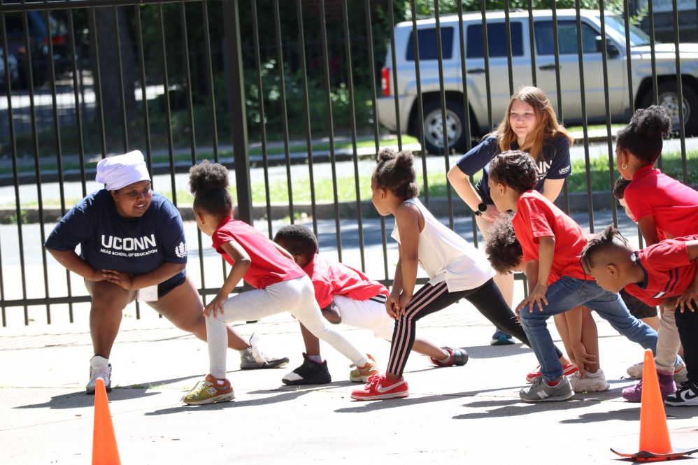 Two college students lead elementary school students in stretching exercises outside.