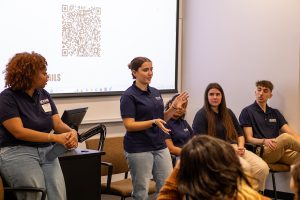 Four college students sit at the front of a classroom while another stands and speaks.