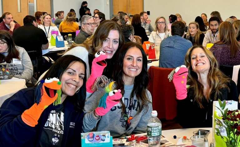 Four women hold up sock puppets with a crowded room behind them.