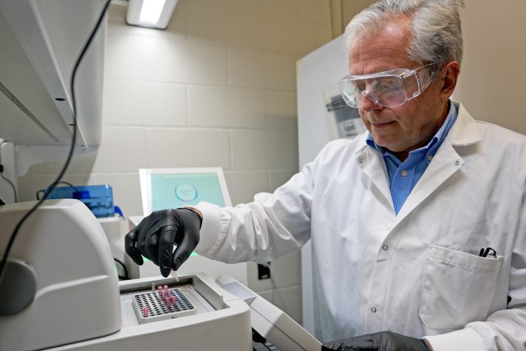 A man wearing a labcoat, goggles, and gloves inserts samples into a machine