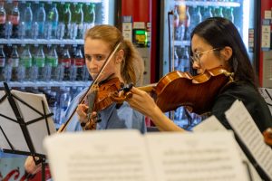 Two women playing violin in front of cafeteria vending machines