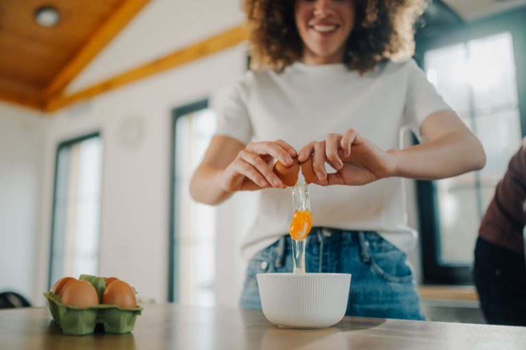 Woman breaking open fresh eggs into white bowl, preparing ingredients for baking