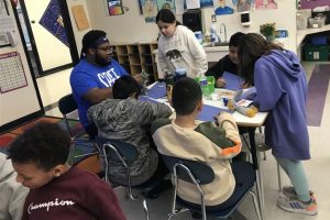 A black college student sits in an elementary school classroom, speaking with five students.