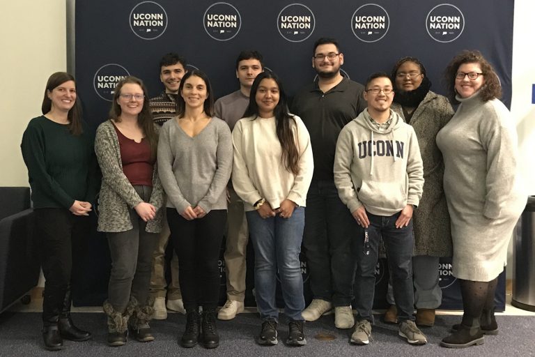 A group of 8 college students and 2 college professors pose in front a navy blue backdrop that reads UConn Nation.