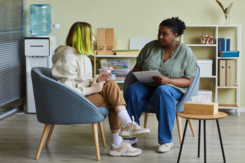 Two coworkers sit across from each other in an office break room, one is younger with dyed green hair.