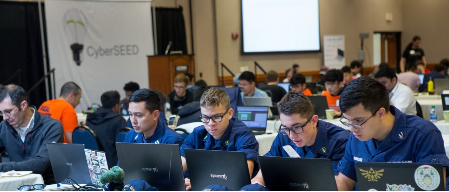 A group of participants in a cybersecurity competition, CyberSEED, working on laptops in a conference hall. Several individuals, some in U.S. Coast Guard uniforms, are focused on their screens, collaborating in teams. The background features a large CyberSEED banner, a podium, and other competitors engaged in the event. The room is filled with tables, chairs, and attendees, creating an atmosphere of intense problem-solving and teamwork.