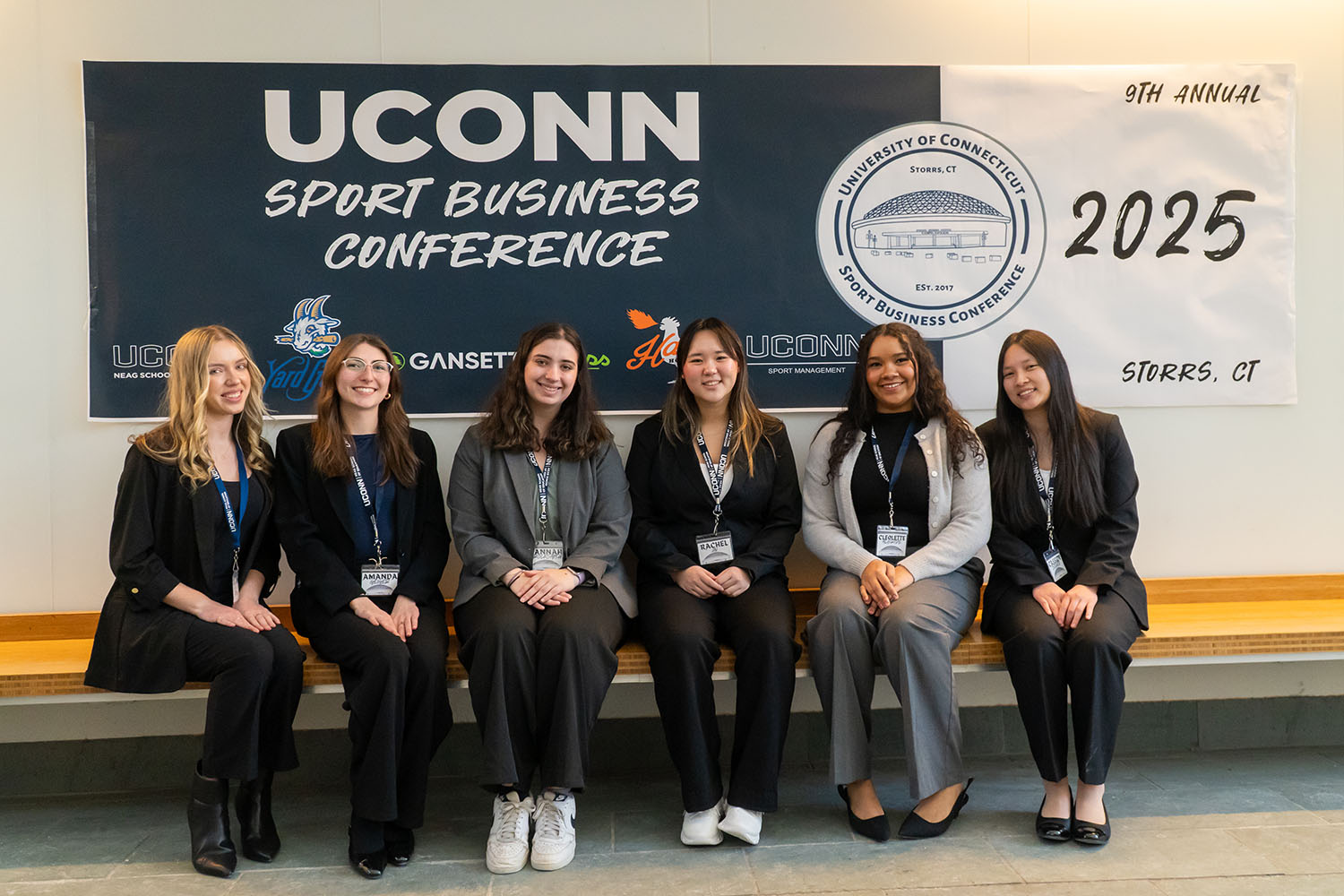 Six female students in professional attire sit on bench in front of the UConn Sport Business Conference banner.