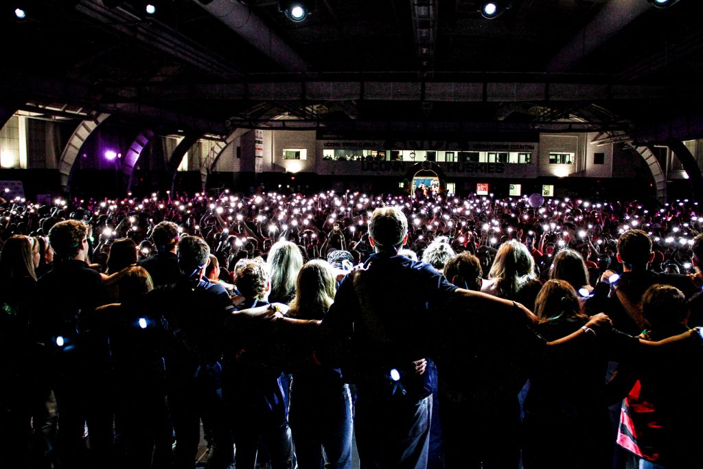 Students link arms at HuskyTHON, looking out at a crowd holding up their phone lights in the darkened field house.