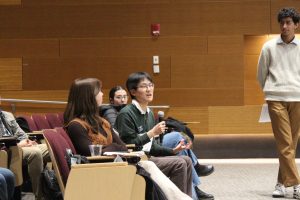 A seated student is holding a microphone and asking a question to a panel of experts.