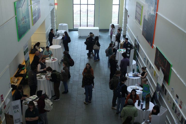 A crowded lobby is full of students talking to groups at tables.