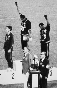Black and white photo with three Olympic Athletes on Podium raising their hands and two men standing near a podium