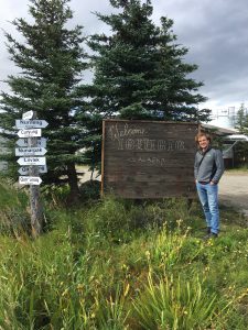 A photo of a person standing next to a wooden welcome sign for a village.