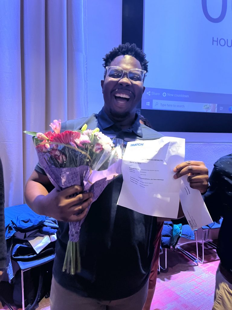 A smiling young man is holding a bouquet of flowers and a piece of paper with writing on it.