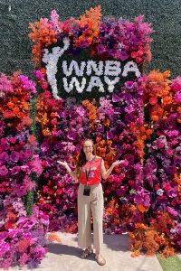 Risa Isard stands in front of a life-size orange and purple floral arrangement with lettering that reads WNBA Way.