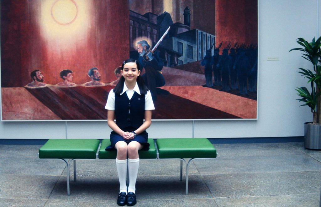 The character Miss Huang from the Apple TV+ show "Severance" sits on a bench in front of a painting.