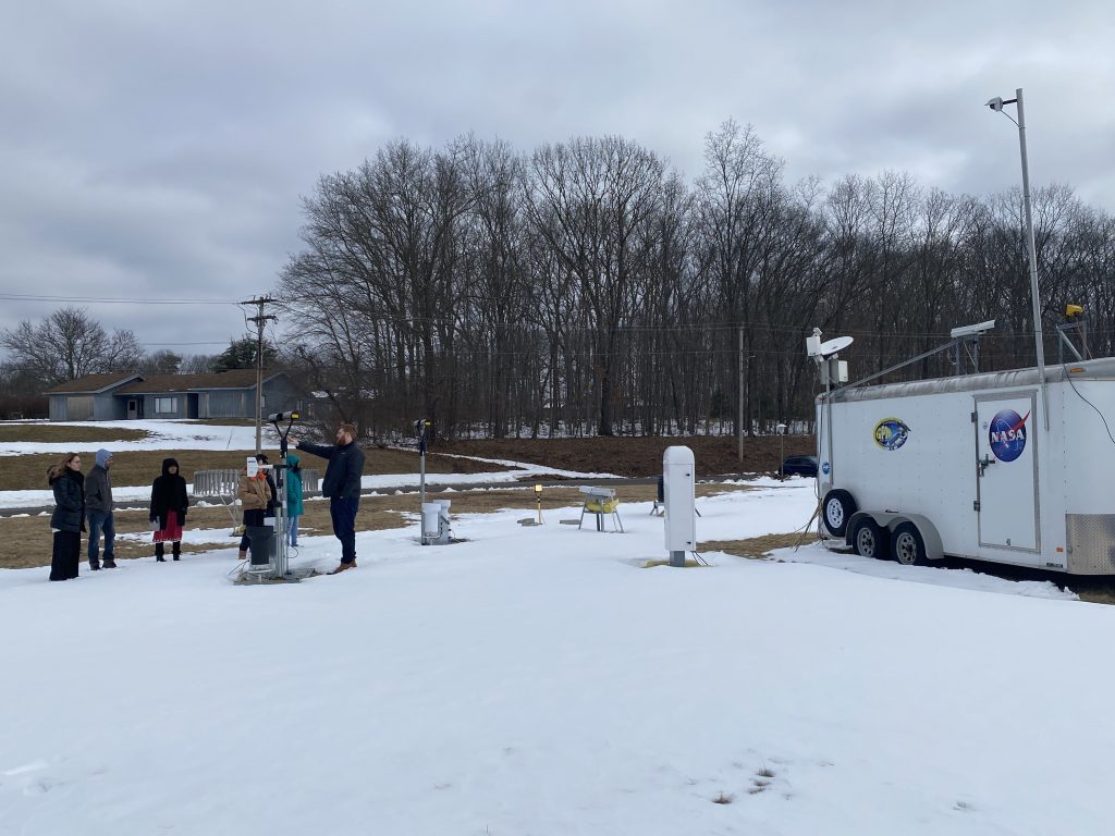 A group of people sand outside in the snow with scientific equipment. A trailer with the NASA logo is off to the side.