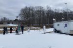 A group of people sand outside in the snow with scientific equipment. A trailer with the NASA logo is off to the side.