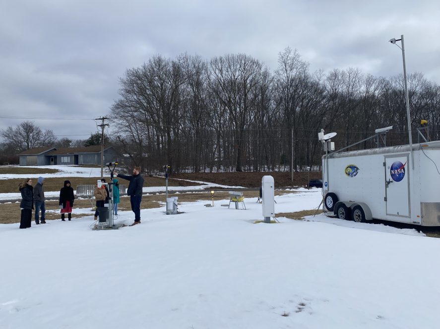 A group of people sand outside in the snow with scientific equipment. A trailer with the NASA logo is off to the side.