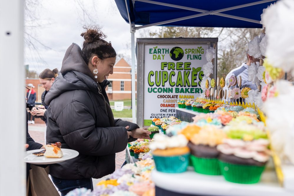 A student in a black jacket selects a cupcake from a table outdoors.