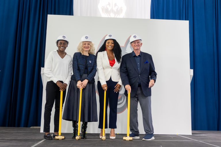 Four people with hard hats and ceremonial sledgehammers stand in front of a white wall.