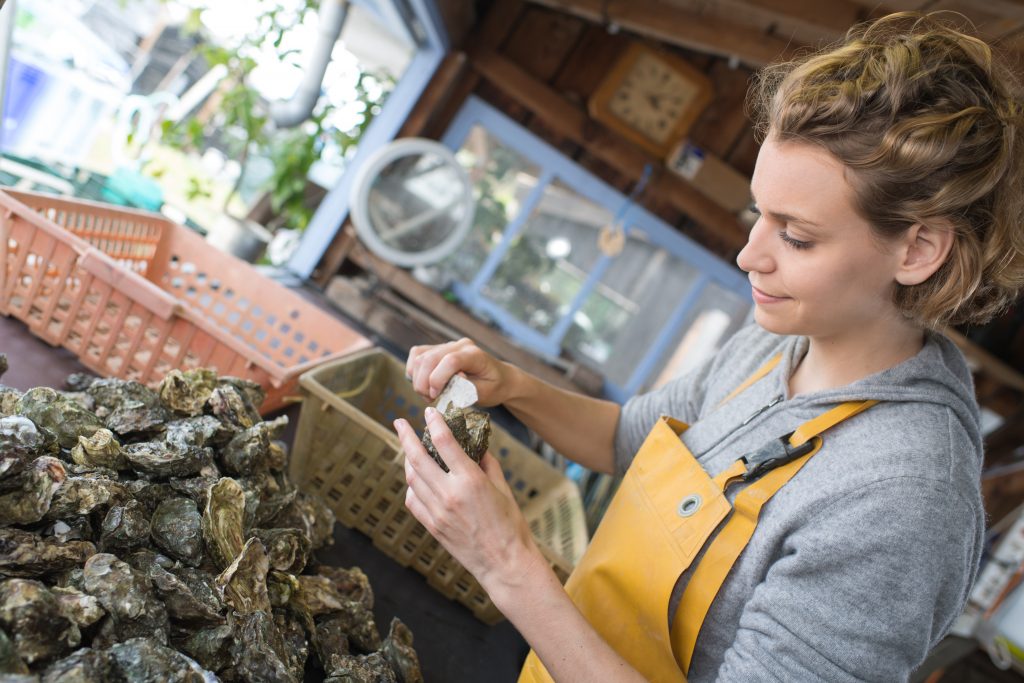A woman in a yellow apron shucks an oyster, with a large pile of shells in front of her.