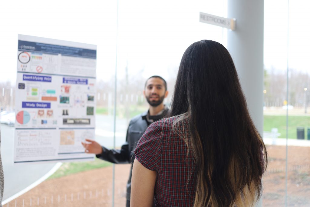 A student describes his research poster, on a window behind him, to a woman whose back is to the camera.