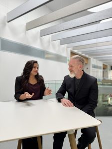 A woman on the left, and a man on the right, both wearing black, sit and talk at a bright white table.