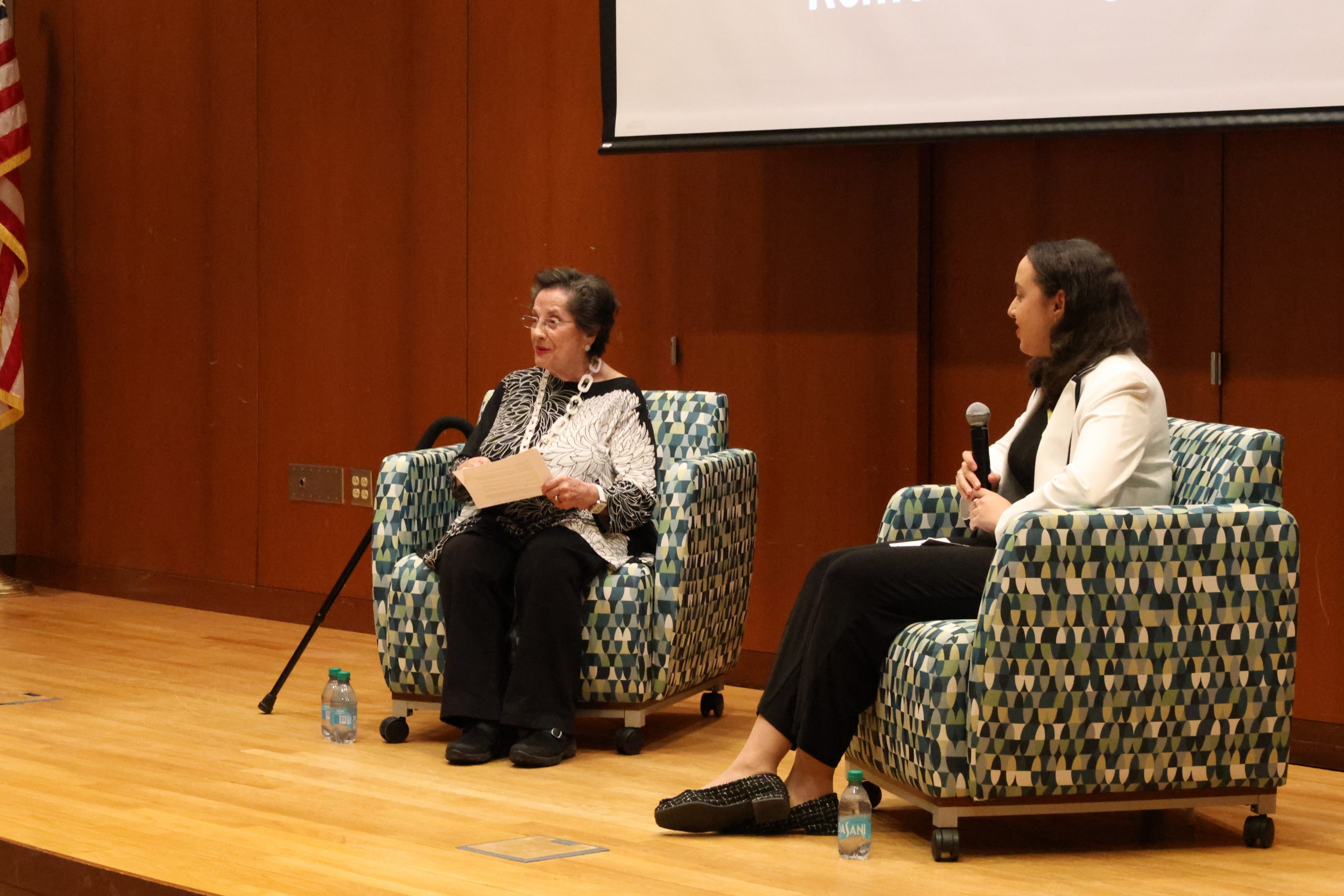 A woman sits on stage in a chair, with a student to her left.