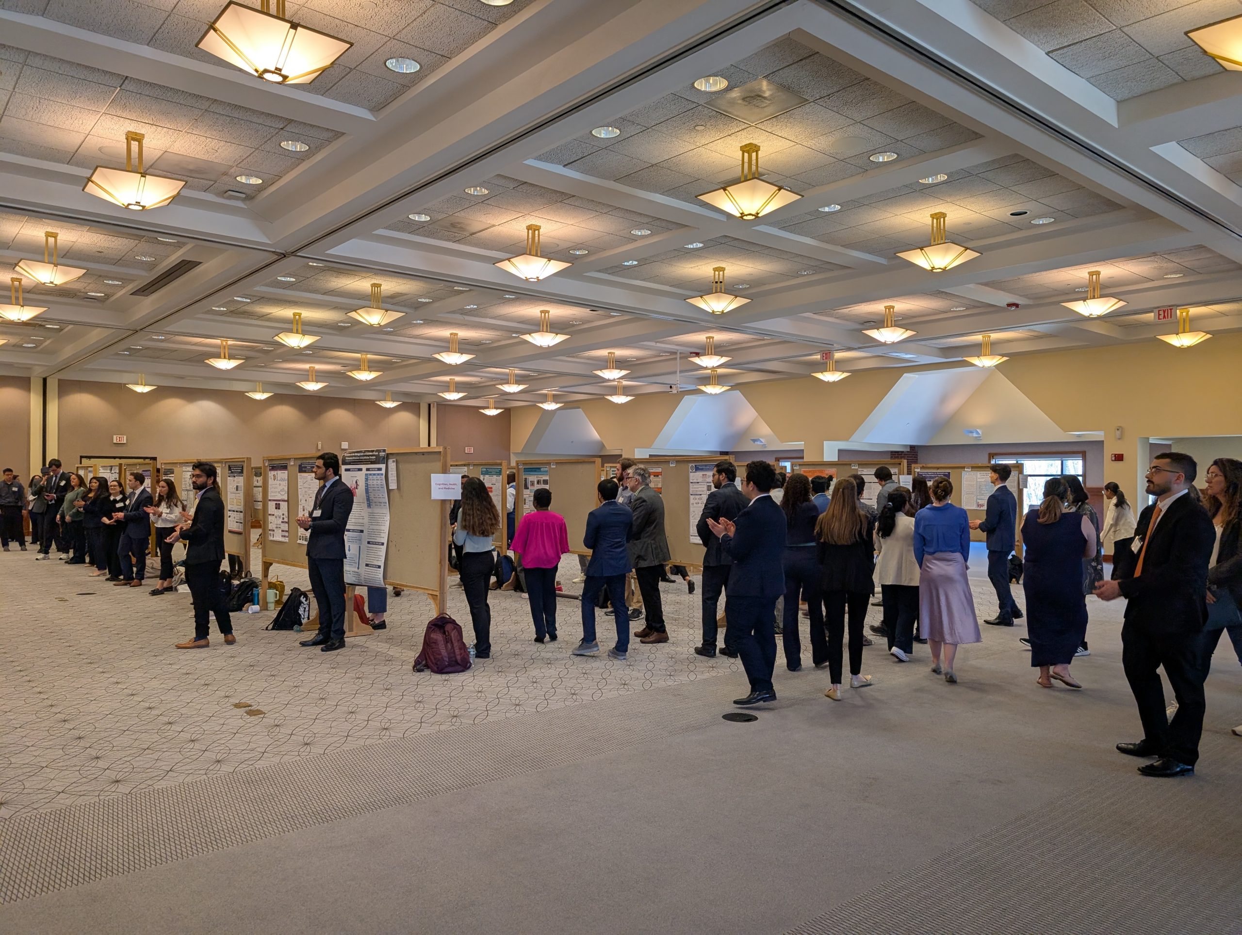 A large group of people standing in a ballroom examine dozens of research posters.