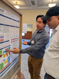 A student in a blue shirt points to a graphic on a research poster while speaking with a student in a white shirt.