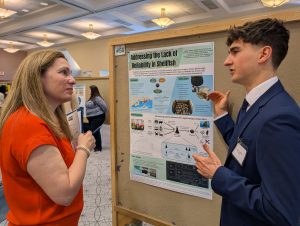 A student in a suit stands before a research poster and explains it to a woman in red.