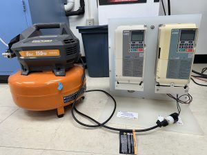 An orange 6-gallon, 150 PSI Ridgid air compressor next to a mounted panel with two Yaskawa A1000 variable frequency drives (VFDs) connected by cables.