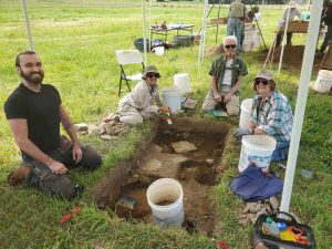 A group of people work under shade tents and dig through green grass into the soil at an excavation site.
